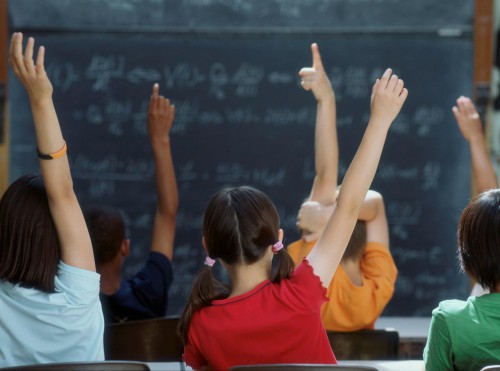 Children sitting in chairs raising their hands in front of a blackboard