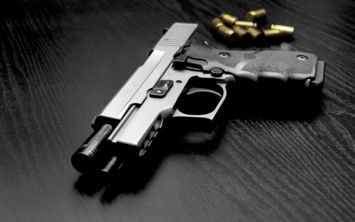 Image of a gun laying on a black table. There are gold bullets in the background