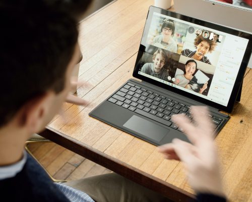 A person sitting at a table facing a laptop with a video meeting screen open with 4 other people on screen.