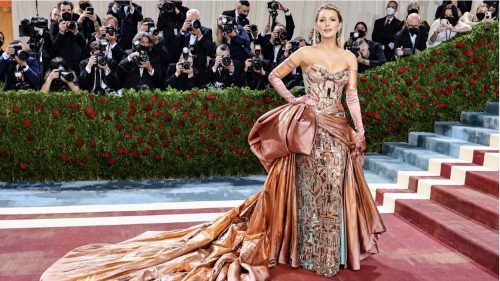 Blake Lively is in a strapless dress with geometric and metallic patterns on the gown. She is wearing metallic colored orange and pink gloves. The dress has a large bow and train also in the metallic orange and pink color. On the dress itself and under the train there is a hint of blue.