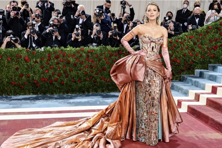 Blake Lively is in a strapless dress with geometric and metallic patterns on the gown. She is wearing metallic colored orange and pink gloves. The dress has a large bow and train also in the metallic orange and pink color. On the dress itself and under the train there is a hint of blue.
